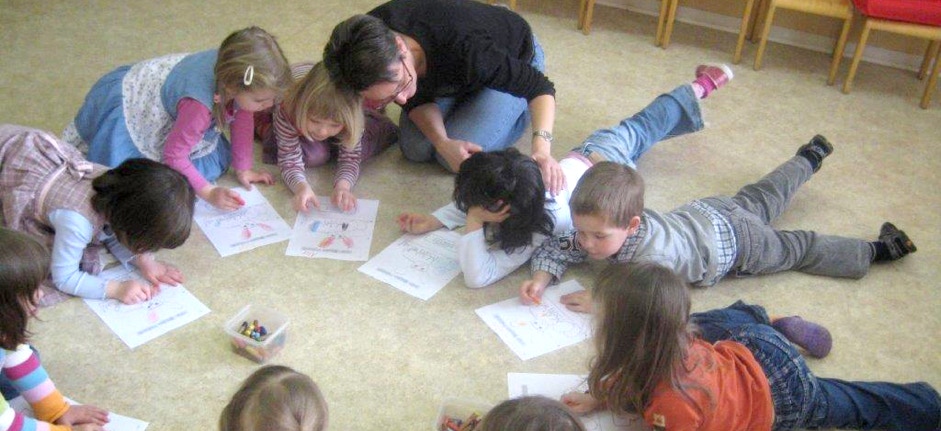Kinder sitzen und lümmeln auf dem Boden und lernen spielerisch Englisch gemeinsam mit einer Lehrerin