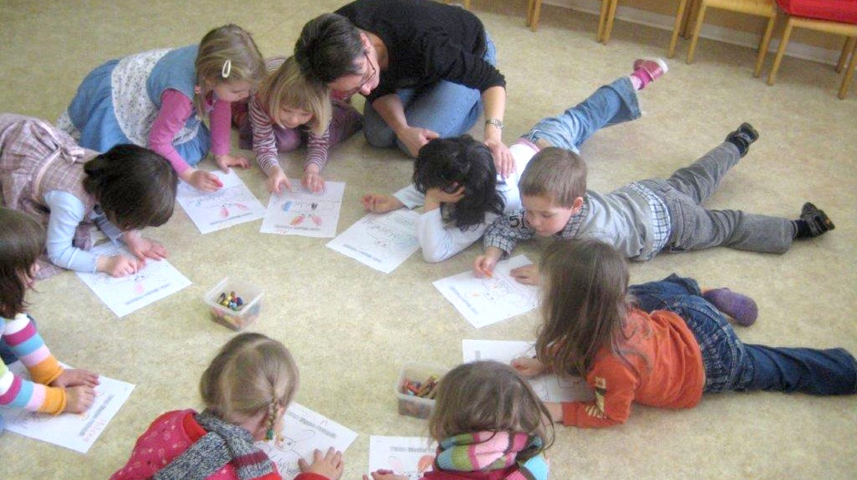 Kinder sitzen und lümmeln auf dem Boden und lernen spielerisch Englisch gemeinsam mit einer Lehrerin