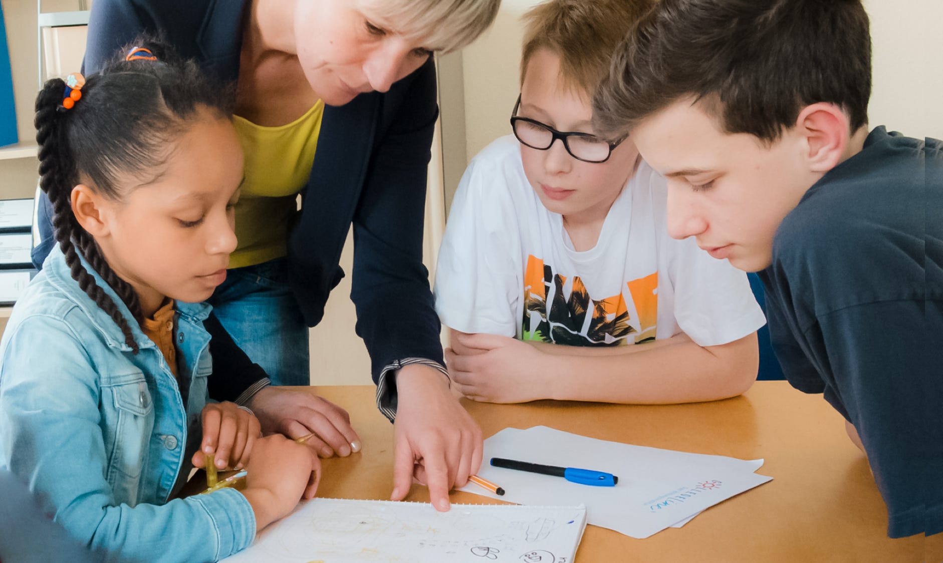 Lehrerin erklärt 3 Schülern eine Aufgabe. Die Kinder hören interessiert zu.