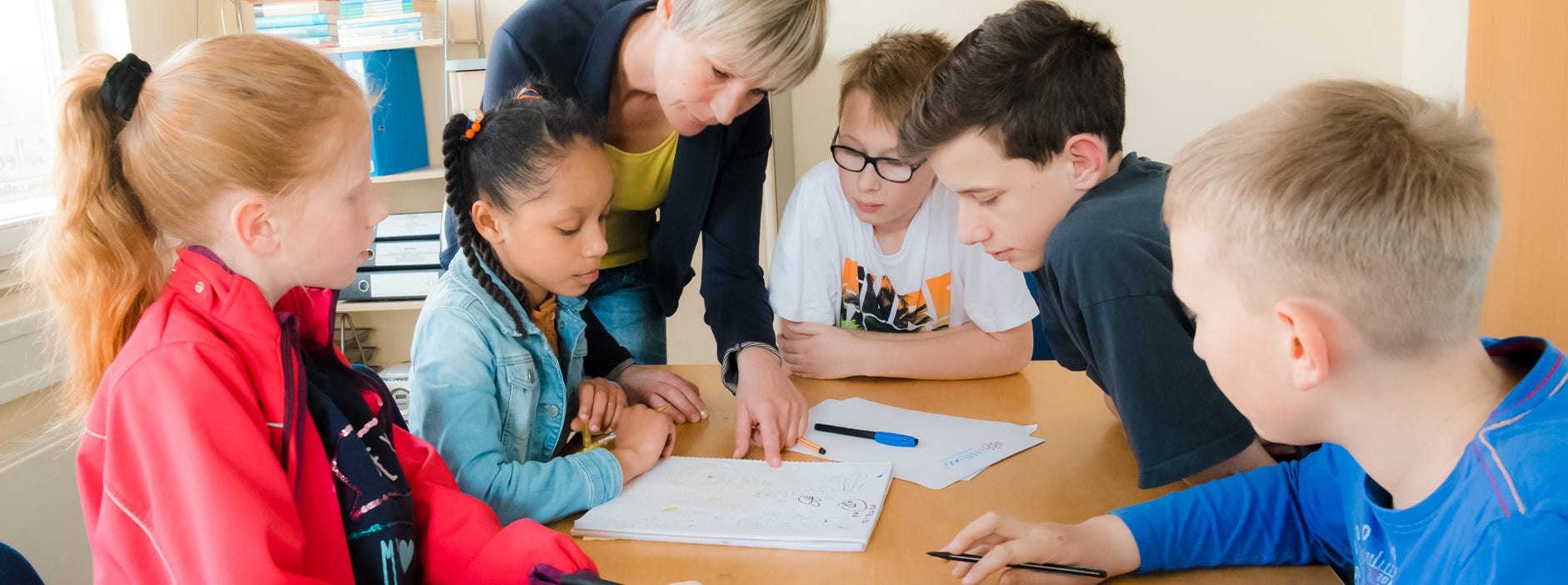 Fünf Schüler hören den Erklärungen der Lehrerin aufmeksam zu. Arbeitsblätter liegen auf dem Tisch.